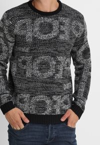 Pull en tricot noir et blanc avec un col rond, présentant une texture à motif audacieux et des poignets et un ourlet côtelés contrastants.