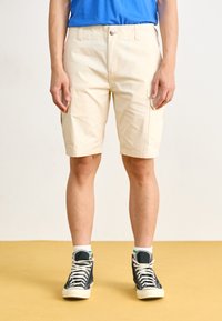 Shorts cargo beige jusqu'aux genoux en tissu léger, dotés de deux poches latérales, d'une fermeture à bouton et d'un motif texturé subtil.