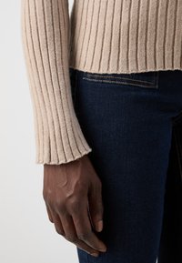 Pull en maille beige à côtes avec des manches longues et un ourlet brut, associé à un jean en denim bleu foncé. Le tissu semble doux avec une finition texturée.