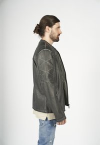 Grå läderjacka med kviltade axelpaneler, dragkedjestängning och en avslappnad passform, bärs över en beige skjorta och slitna blå jeans.