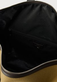 Ανοιχτή υφασμάτινη τσάντα σε ανοιχτό καφέ χρώμα με ασημένιο φερμουάρ που δείχνει την μαύρη εσωτερική επένδυση και μια εσωτερική τσέπη με φερμουάρ, με την ετικέτα Boss.