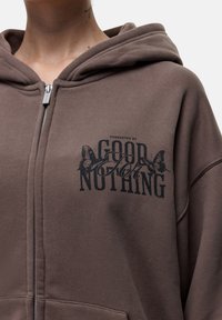Felpa con zip marrone con una grande scritta grafica nera che recita "GOOD NOTHINGS" e accenti a forma di farfalla sulla parte anteriore. Tessuto morbido e texturizzato.