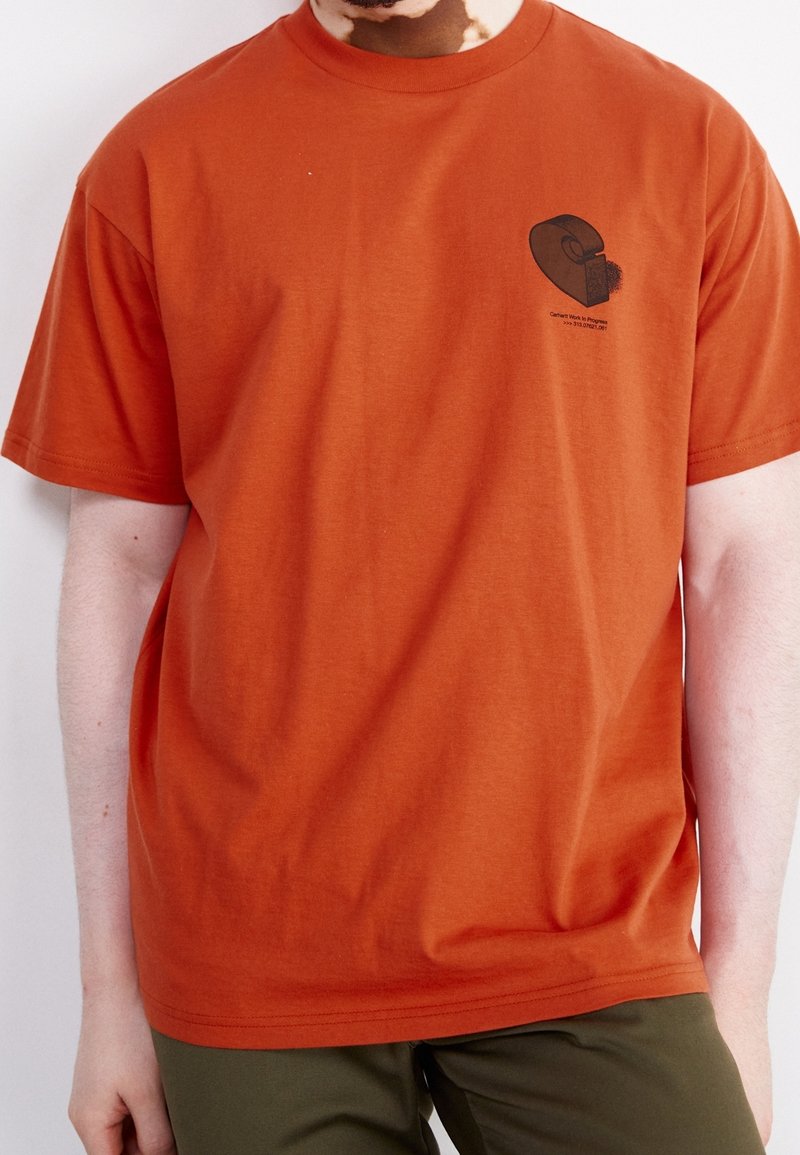 Hombre que lleva una camiseta naranja quemado con un pequeño logotipo abstracto negro y texto en la parte superior izquierda del pecho, combinada con pantalones verdes oliva.