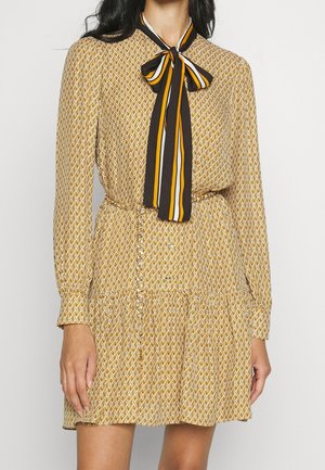 Kobieta w beżowej sukience z długim rękawem we wzór łańcuszków oraz z czarną, białą i żółtą kokardką w paski przy kołnierzu.