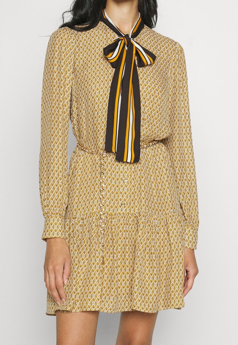 Femme portant une robe beige à manches longues avec un motif à maillons de chaîne et un nœud papillon rayé noir, blanc et jaune au col.