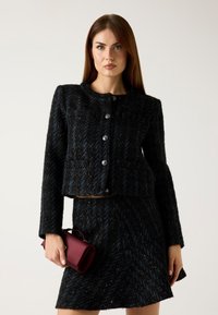 Giacca in tweed nero con dettagli blu, scollo rotondo e chiusura con bottoni; abbinata a una gonna coordinata e a una pochette bordeaux.