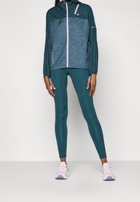Teal let jakke med tekstureret mønster, lyserød lynlås og hætte; parret med ensfarvede teal leggings og hvide sneakers med lilla detaljer.