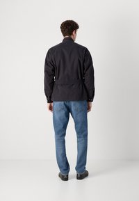 Casaco azul-marinho de mangas compridas com cintura cintada e punhos com botões, acompanhado por jeans azul-claro e sapatos pretos, visto pela parte de trás.
