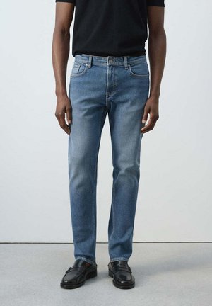 Mann trägt mittelblaue Jeans mit geradem Bein, Vordertaschen und Knopfverschluss, kombiniert mit schwarzen Loafers und einem schwarzen Hemd.