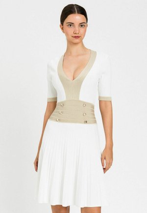 Donna che indossa un vestito plissettato bianco con scollo a V profondo beige e ampia fascia in vita con sei bottoni, in piedi davanti a uno sfondo chiaro e uniforme.