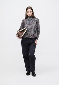 Szürkés mintás blúz fodros részlettel, sötét texturált nadrág és fekete cipő. Barna és krémszínű clutch táskát tart a kezében.