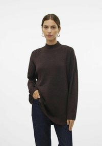 Pull en tricot marron avec un col haut, coupe loose, poignets côtelés et fentes sur les côtés. Porté avec un pantalon en denim foncé, présentant une texture lisse.
