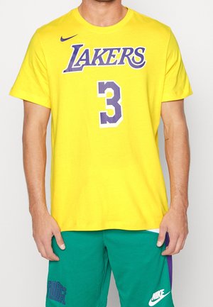 Mężczyzna w żółtej koszulce Lakers z numerem 3 oraz zielonych krótkich spodenkach Nike z niebieskimi i białymi logo, stoi z rękami swobodnie opuszczonymi wzdłuż ciała.