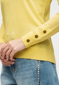 Personne portant un pull jaune à manches longues avec trois boutons marron sur le poignet et un jean bleu avec des clous métalliques sur la couture latérale.