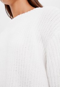 Närbild av en person som bär en vit tjock stickad tröja med rund halsringning och texturerat mönster.