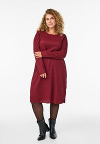 Femme aux cheveux bouclés portant une robe bordeaux à manches longues avec de petits pois, des collants noirs et des bottines noires, se tenant les bras croisés.