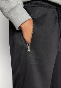 Schwarze Jogginghose aus glattem Stoff mit einer sichtbaren Reißverschlusstasche und elastischem Bund; ergänzt durch einen hellgrauen Sweatshirtbund.