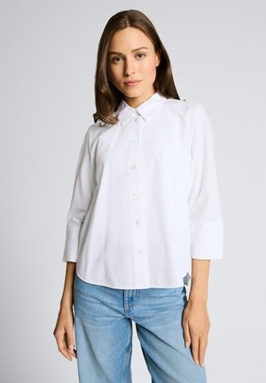 Młoda kobieta ubrana w białą koszulę zapinaną na guziki z rękawami do łokci i jasnoniebieskie dżinsy, stojąca na tle jednolitego szarego tła.