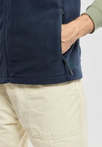 Gilet en micropolaire marine avec une fermeture éclair et des poches latérales, présentant une texture lisse. Assorti à un pantalon décontracté de couleur claire.
