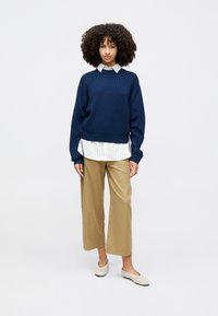 Suéter de malha azul marinho por cima de uma camisa branca de colarinho, combinado com calças largas bege e sapatos claros. Design e ajuste simples, com laços laterais.