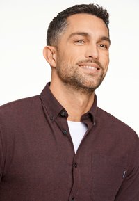 Viininpunainen napitettava paita, joka on valmistettu teksturoidusta kankaasta. Paidassa on rintatasku ja mustat napit, ja se on kerroksittain valkoisen pyöreäkauluksisen t-paitan päällä.