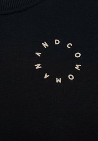 Zwarte sweatshirt met een cirkelvormig ontwerp in beige, met de woorden "AND COMPANY" en "MAN." De tekst heeft een casual, ongelijkmatige stijl.