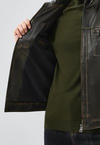 Veste en cuir noir avec une texture lisse, fermeture éclair et poche intérieure. Portée par-dessus un haut en tricot vert, mettant en valeur un design ajusté.