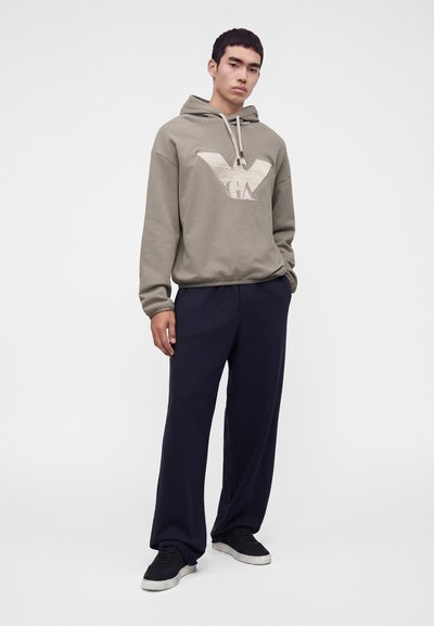 Jeune homme portant un sweat à capuche gris avec un grand logo, un pantalon large bleu marine et des baskets noires, debout avec une main dans la poche.