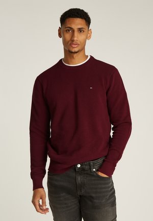 Junger Mann, der einen dunkelroten Langarmpullover und schwarze Jeans trägt und mit einer Hand in der Tasche vor einem einfarbigen Hintergrund steht.