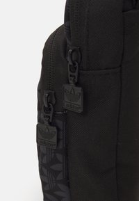 Mochila de tela negra con un costado estampado, que cuenta con dos cremalleras negras y tiradores de cremallera con la marca. Material texturizado con detalles de diseño sutiles.