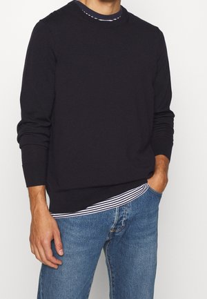 Pull en maille navy à col rond avec des poignets côtelés, superposé sur un T-shirt à rayures blanches et navy, associé à un jean en denim bleu.