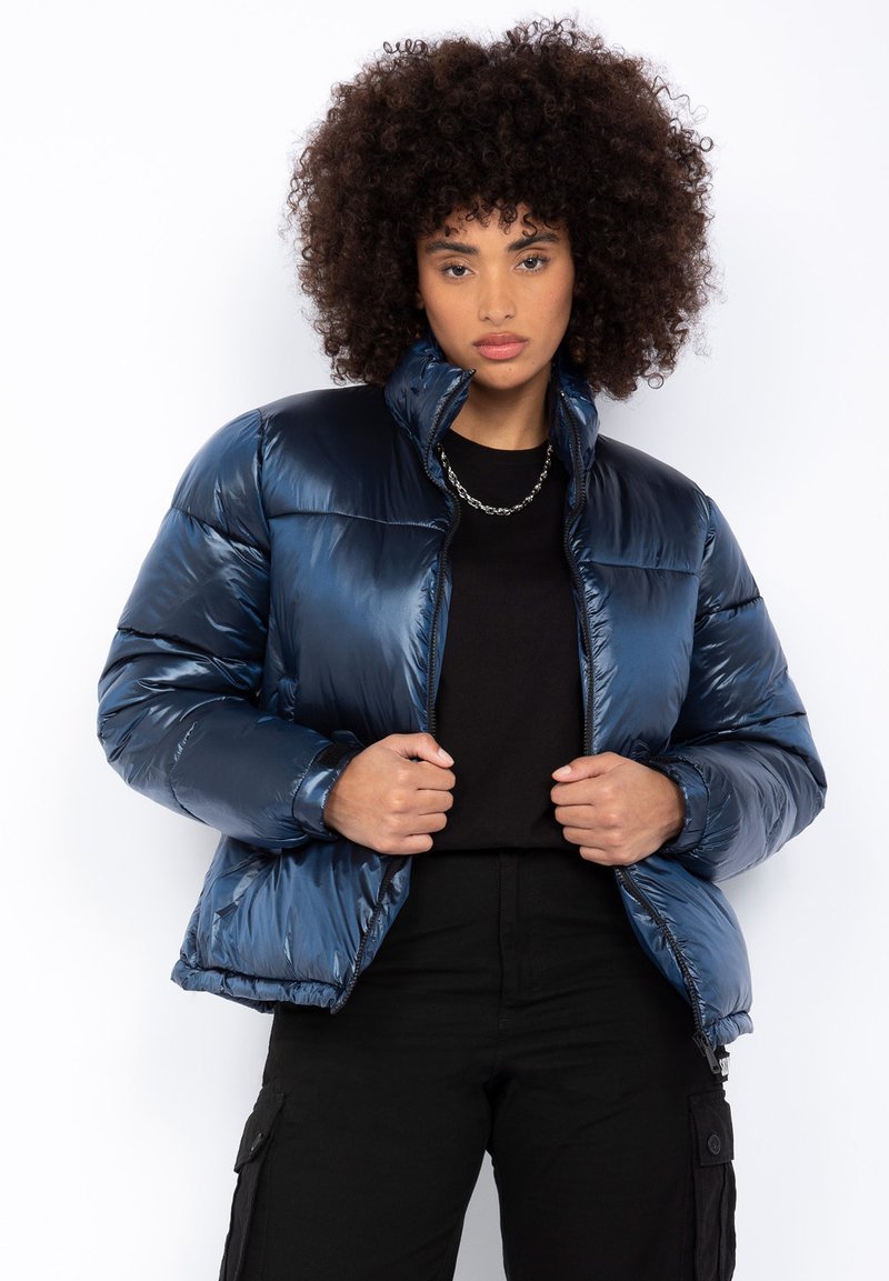 Veste bleu marine rembourrée avec col haut, texture brillante et fermeture zippée. Présente une silhouette carrée et des poignets élastiques.