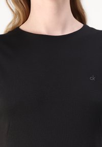 Crna majica s crewneck izrezom od mekog materijala, s diskretnim izvezenim "ck" logom na lijevoj strani. Jednostavan, uski dizajn.