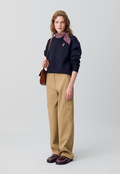 Sudadera de marina con mangas acampanadas, pantalones cargo anchos de caqui con bolsillos y mocasines marrones con acentos plateados. Bufanda a cuadros rojos y blancos.