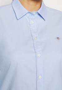 Himmelblaues Hemd mit Knöpfen und Kragen, mit weißen Knöpfen und einem kleinen bestickten Logo auf der Brust. Glatte Textur.