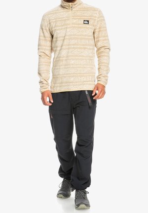 Uomo che indossa un pullover beige con motivo e zip fino a metà, pantaloni cargo neri con zip e scarpe da trekking grigie, in piedi su sfondo bianco.