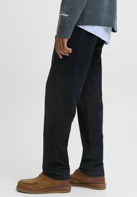 Pantalons en velours côtelé noirs avec une coupe droite, présentant de subtiles rayures verticales. Portés avec un pull en maille gris et des chaussures en daim marron.