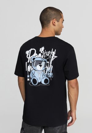 T-shirt nera in cotone con una grande grafica blu di un orsetto che indossa occhiali da sole sul retro, con testo e accenti in stile graffiti.
