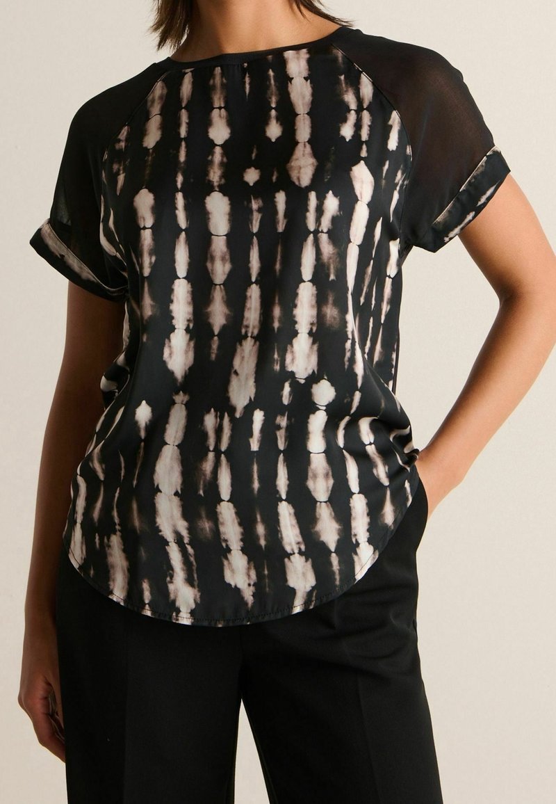 Femme portant un t-shirt noir à manches courtes avec des motifs tie-dye verticaux beiges, associé à un pantalon noir uni, debout avec une main dans la poche.