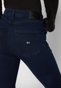 Mørkeblå jeans med en tettsittende design, med en svart lærmidjeetikett og to baklommer med en liten merkelapp.