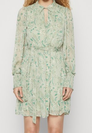 Femme portant une robe vert clair à motif cachemire, manches longues, poignets smockés, ceinture à la taille et encolure en trou de serrure avec un nœud à l'avant.