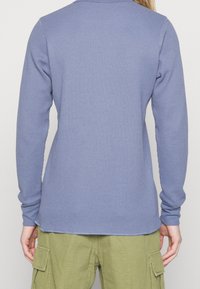 Personne portant une chemise en bleu texturé à manches longues et un pantalon cargo vert olive, vue de dos avec les bras détendus le long du corps.