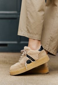 Beige fuzzy sneakers with a black leather accent, thick rubber sole, and laces. Textured surface with branding near the heel.
