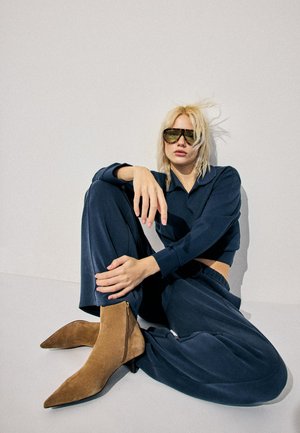 Donna con capelli biondi e grandi occhiali da sole seduta contro una parete bianca, indossa un completo blu navy e stivaletti alla caviglia in camoscio color cuoio con punta appuntita.