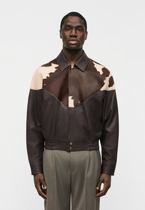 Homme portant une veste en cuir à motif peau de vache marron et beige avec un pantalon de costume beige, debout devant un fond clair uni.