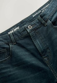 Blugi din denim de culoare albastru închis, cu o croială ajustată, având închidere cu nasturi, bucle pentru curea și eticheta "Denim Supply" la talie.