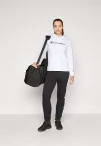 Sweat-shirt à capuche blanche avec le logo "Champion" noir, associé à un pantalon de survêtement noir, tenant un sac de sport noir, sur un fond uni.