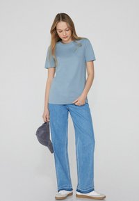 Světle modré tričko s kulatým výstřihem vyrobené z bavlny, spárované s světle modrými širokými džínami. Doplněk zahrnuje šedou kšiltovku drženou v levé ruce.