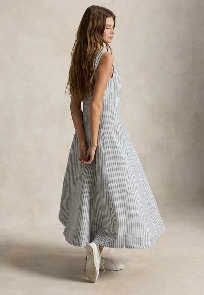 Γυναίκα που φοράει ένα μαν sleeveless, φόρεμα ρίγες μέχρι τον αστράγαλο και λευκά αθλητικά παπούτσια, πόζα με τα χέρια πίσω από την πλάτη της πάνω σε ένα μονόχρωμο φόντο.