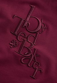 Burgundská bavlnená tkanina s vyvýšeným vyšívaným textom "by Ted Baker" a malým detailom štvorlístka. Hladká textúra.